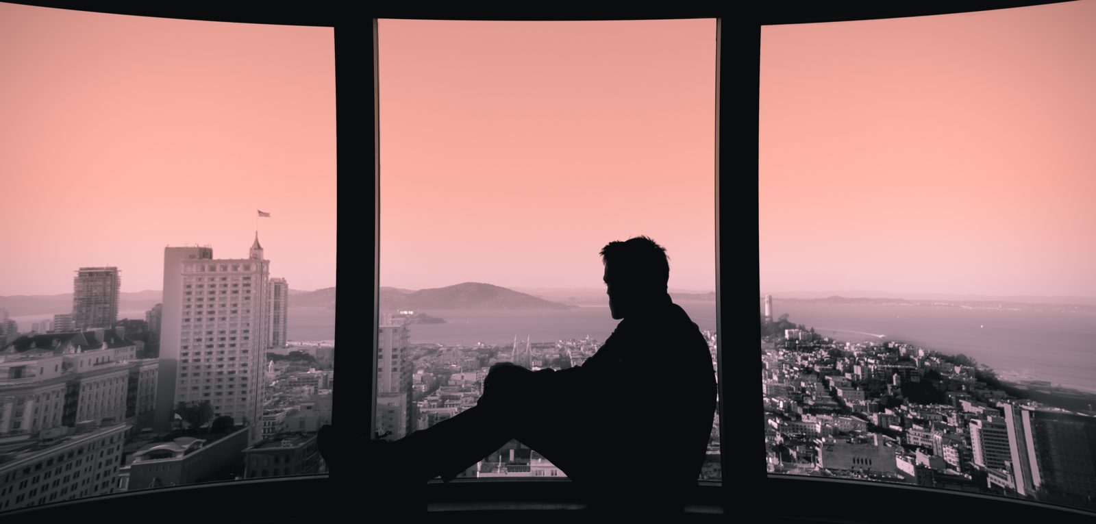 A lone figure seated at a wide window, looking out over a city at dusk